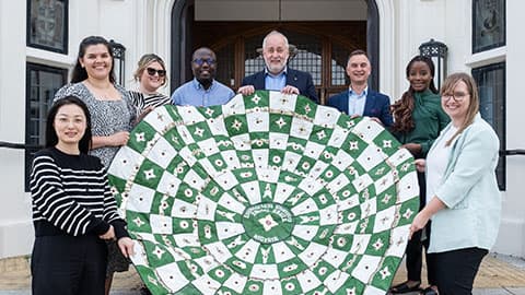 Loughborough University staff from the Vice-Chancellor's Office, Alumni Relations Team and Global Engagement Team holding a tapestry made by alumni in Abuja  in front of Hazlerigg Building on Loughborough University campus. They are all smiling in the sunshine and standing on the steps outside the doorway.